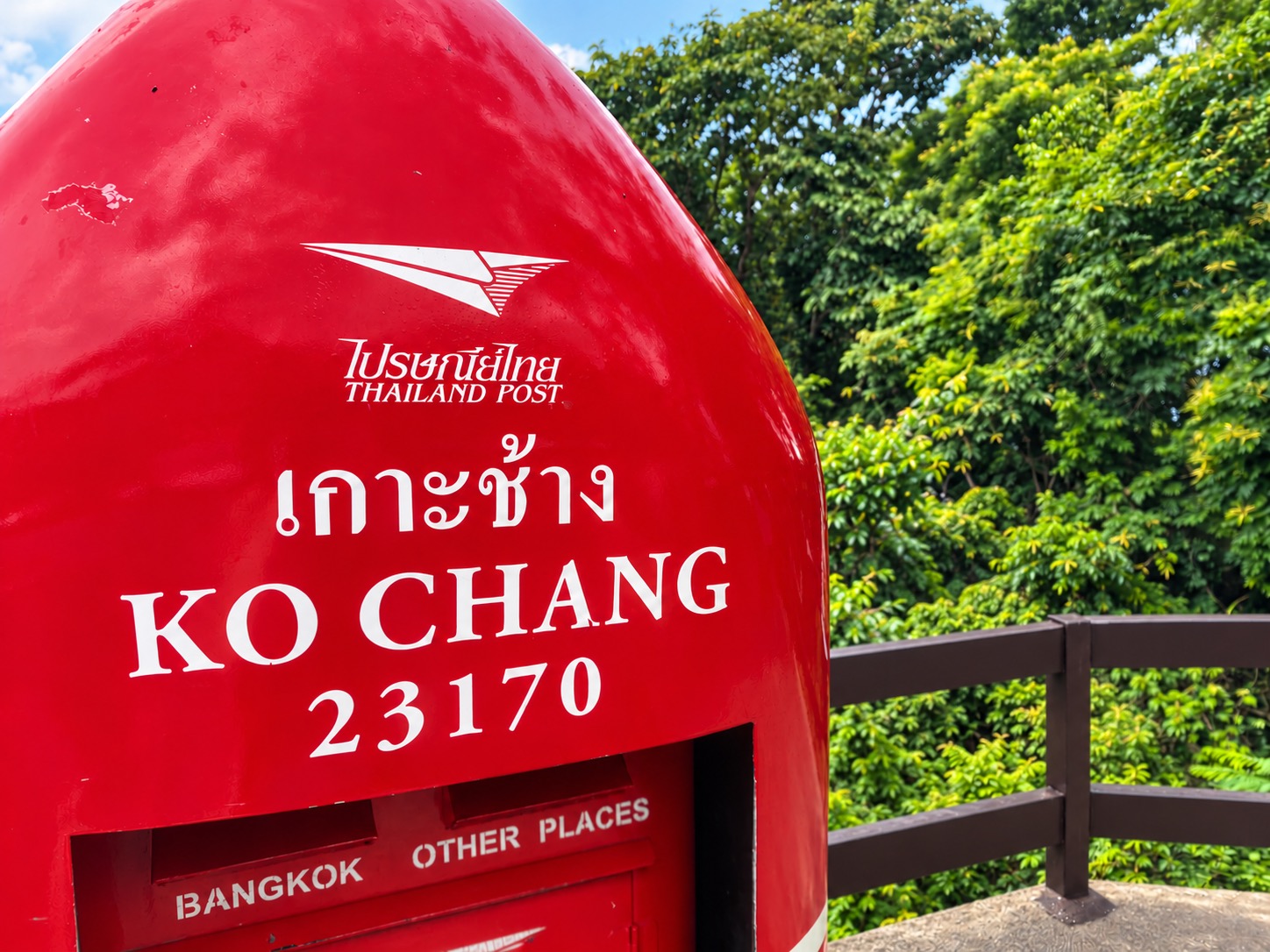 Large red Ko Chang postbox beside a wooden railing and trees